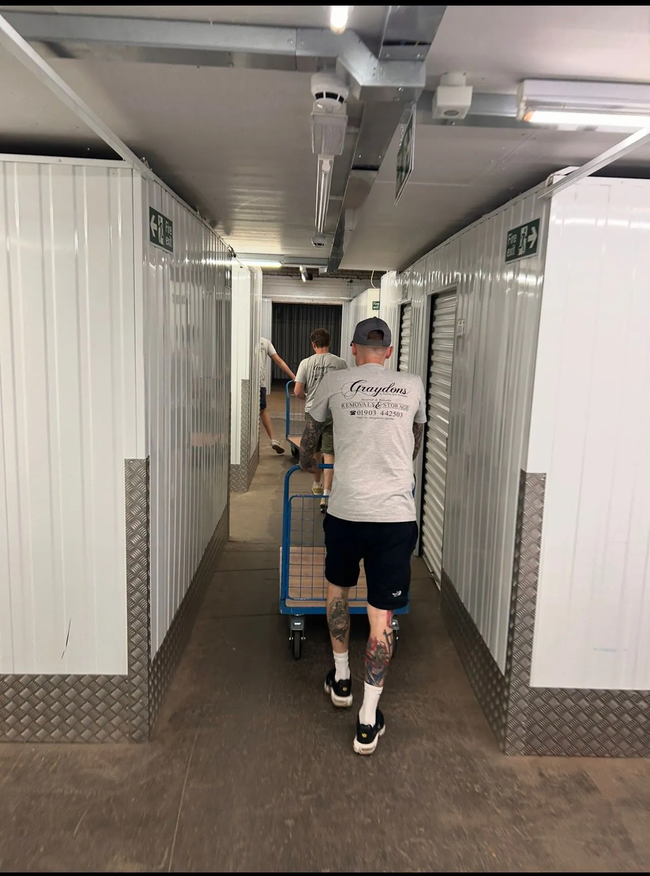 Two movers in matching shirts pushing empty trolleys down a storage unit corridor.