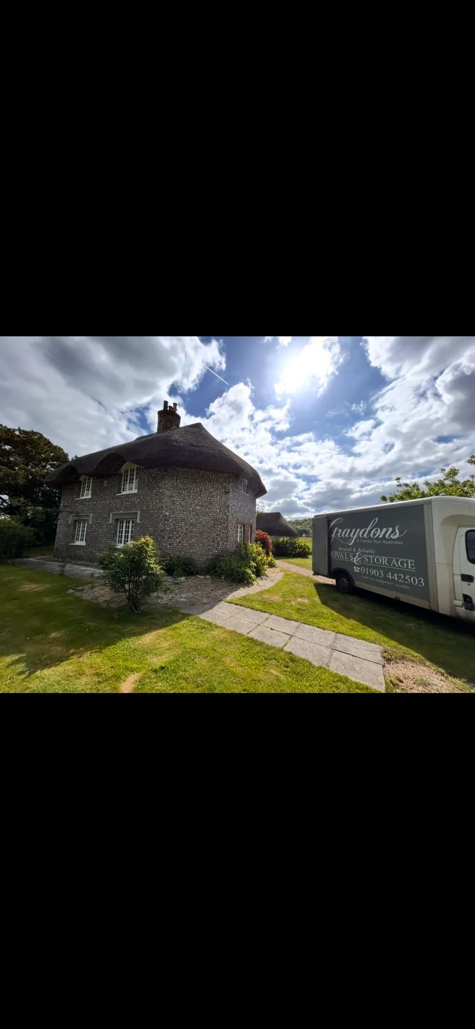 Stone cottage with thatched roof and chimney in a sunny garden with a storage van nearby.