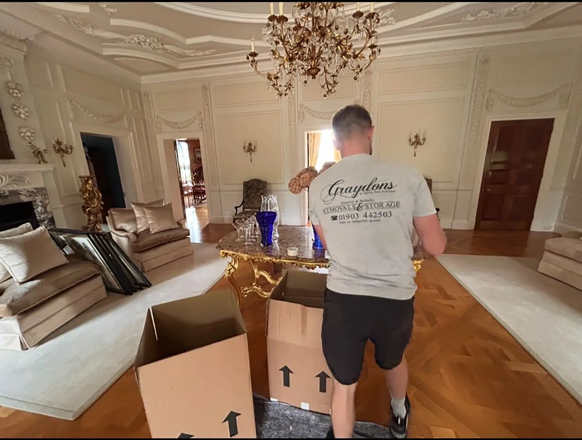 Man wearing Graydons Removals & Storage shirt packing cardboard boxes in an ornate living room.
