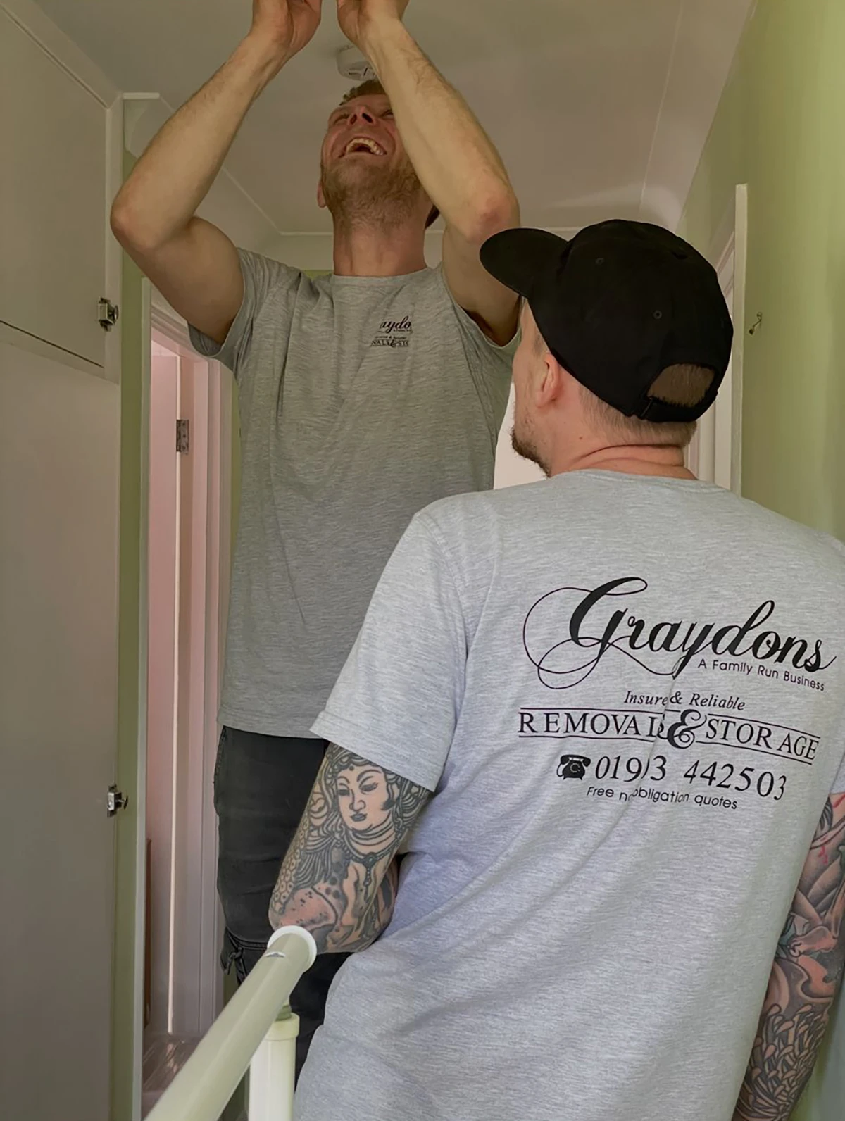 Two removal men in gray shirts working on a ceiling light fixture in a hallway.