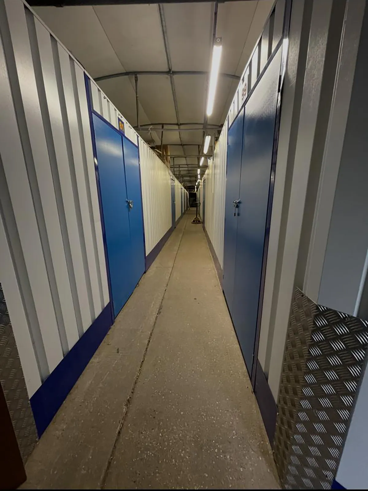 Long narrow corridor between white storage units with blue doors and concrete floor under fluorescent lights.