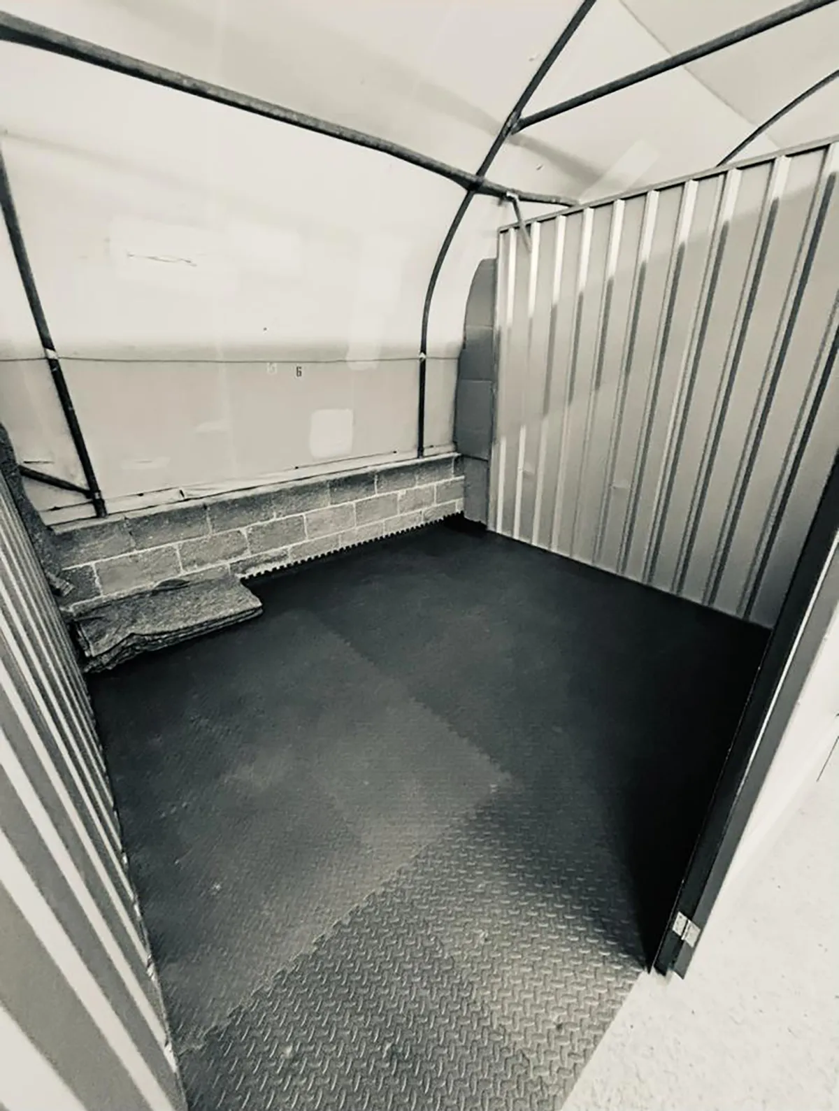 Empty small storage unit with black rubber flooring and corrugated metal walls inside.