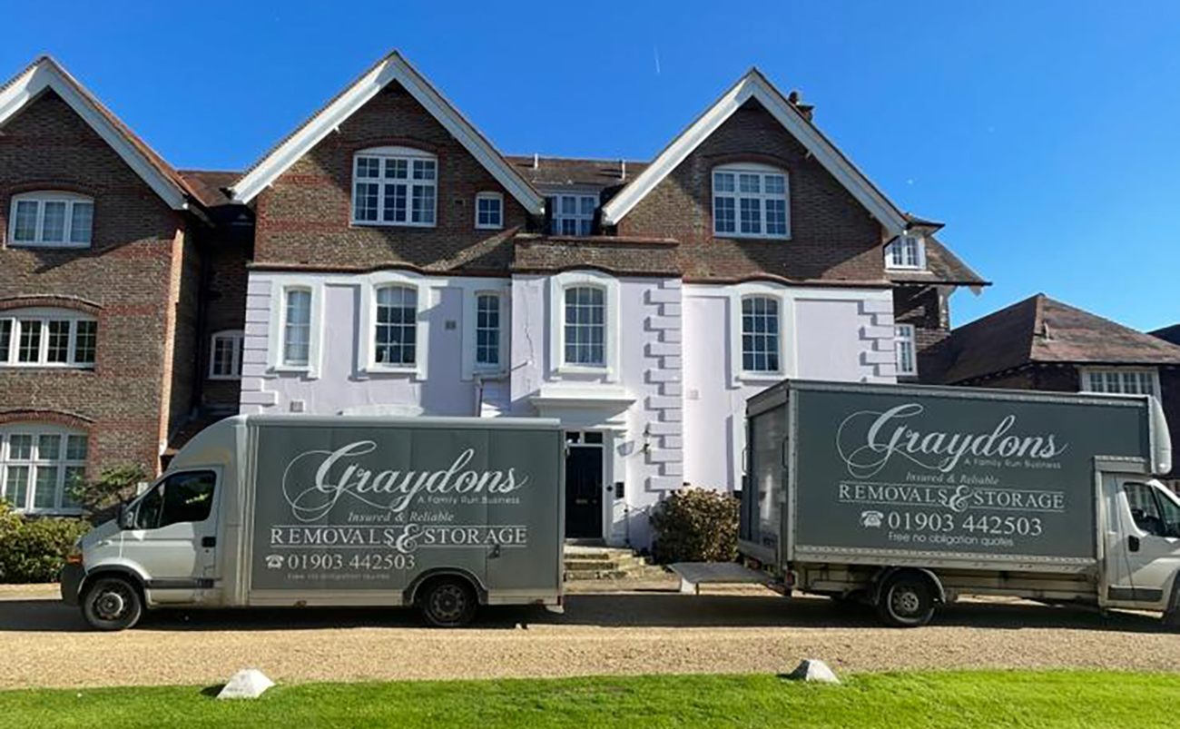 Two Graydons removals and storage trucks parked outside a large brick and white house under a clear sky.