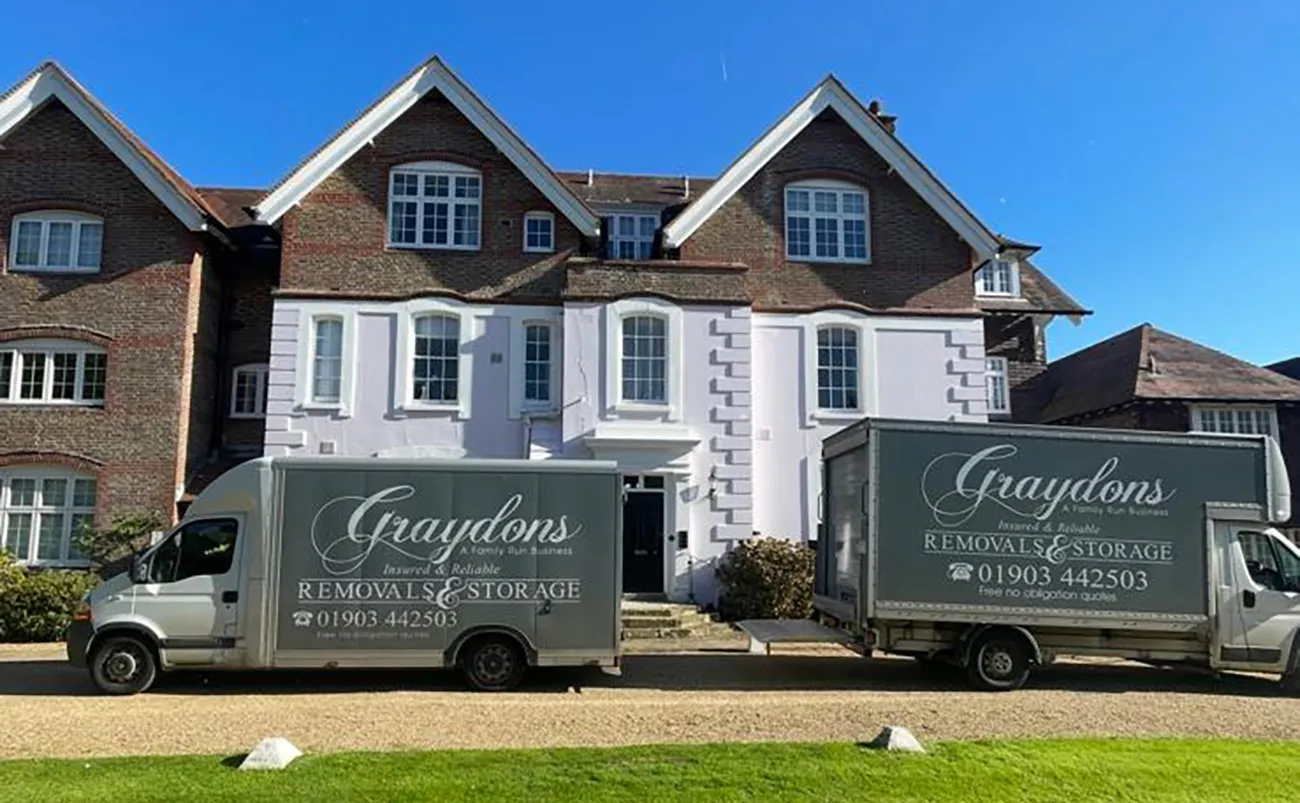 Two Graydons removals and storage trucks parked outside a large brick and white house under a clear sky.