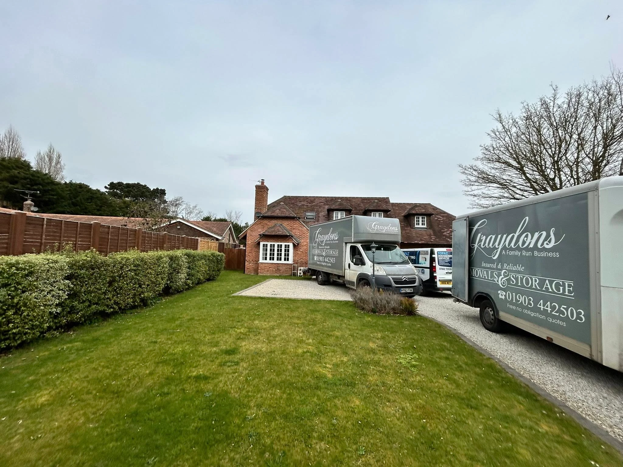 Three Graydons removals and storage vans parked on driveway of brick house with green lawn and hedge.