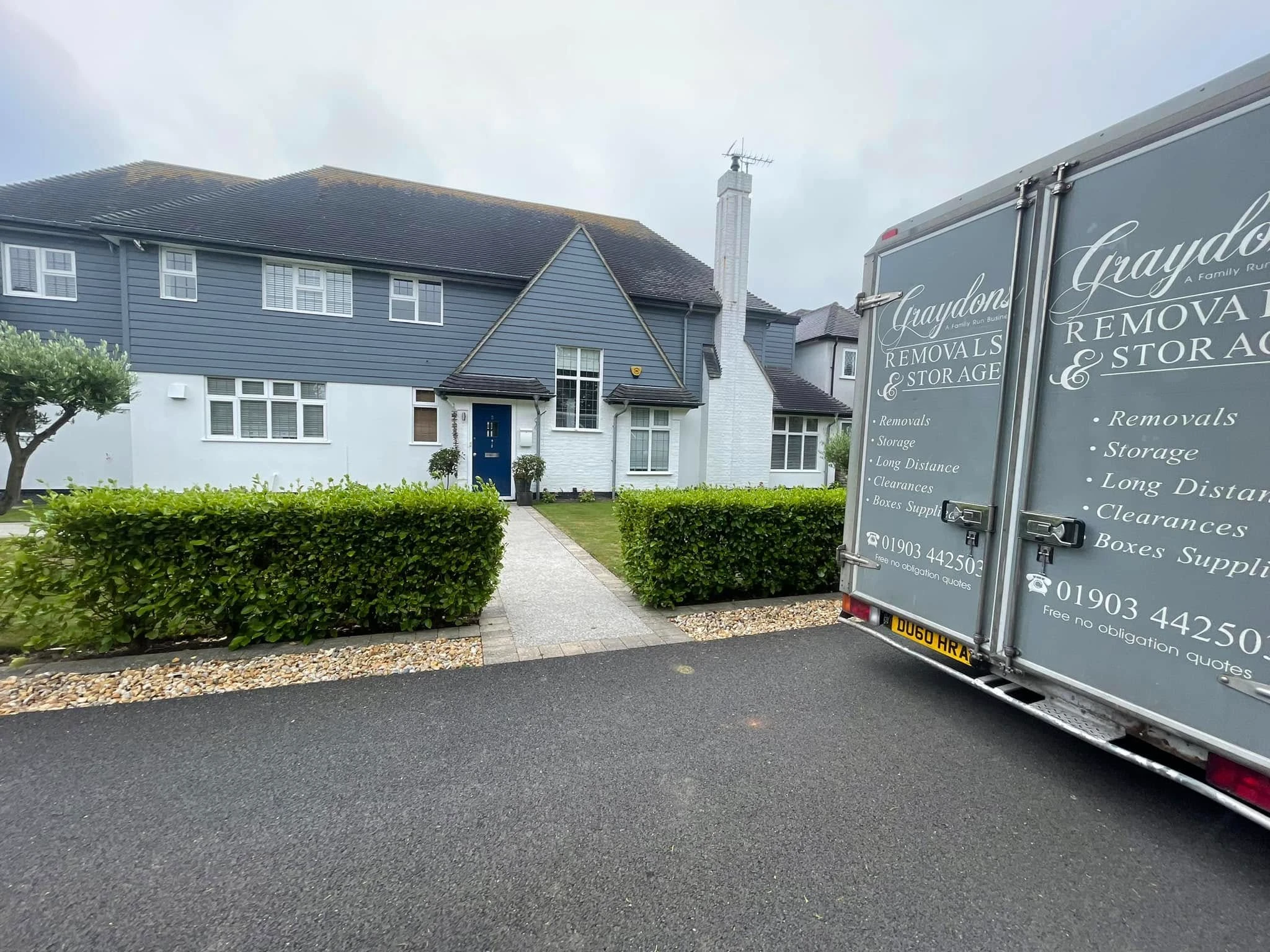 Gray and white two-story house with blue door, shrubs, and Graydons Removals truck in driveway.