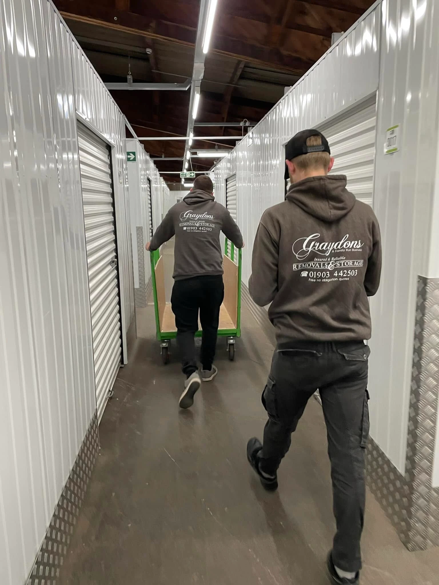 Two men in Graydons hoodies moving an empty green cart down a warehouse hallway.