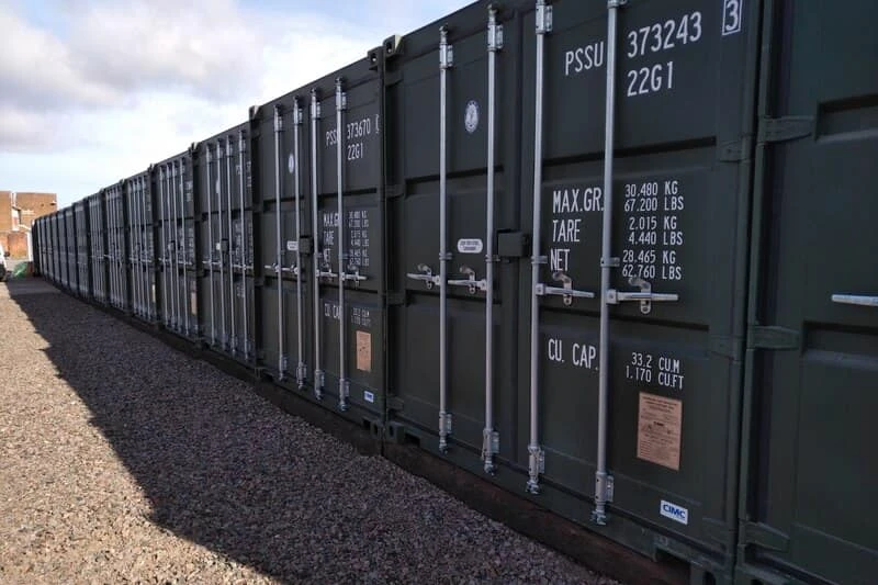 Long row of dark shipping containers with visible white weight and capacity markings on gravel ground.